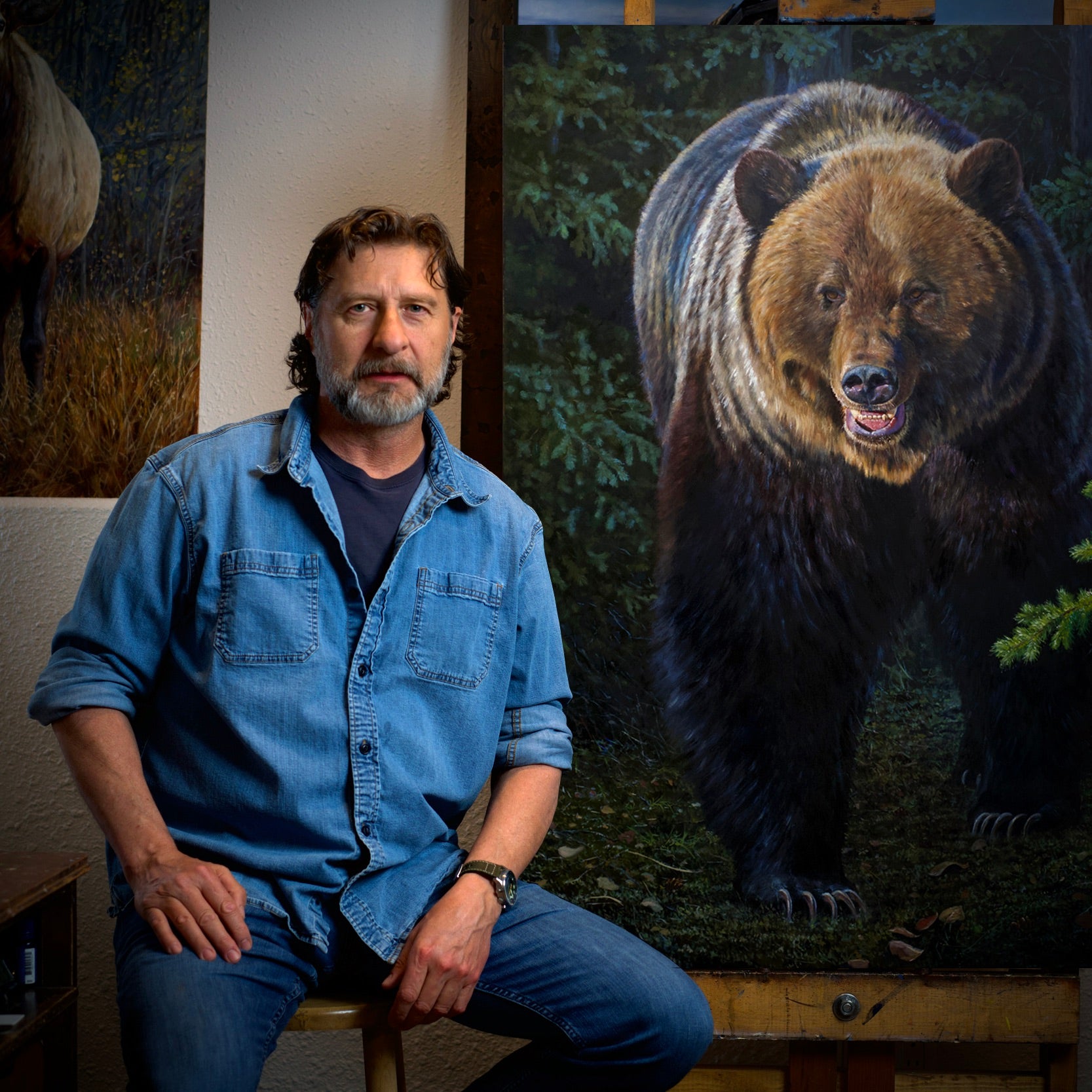 “Studio portrait of wildlife artist with his original oil painting ‘What Lurks Within,’ featuring a powerful grizzly bear stepping out from the forest shadows. This image represents the artist’s deep connection to wildlife and the dramatic realism of his painting style.”