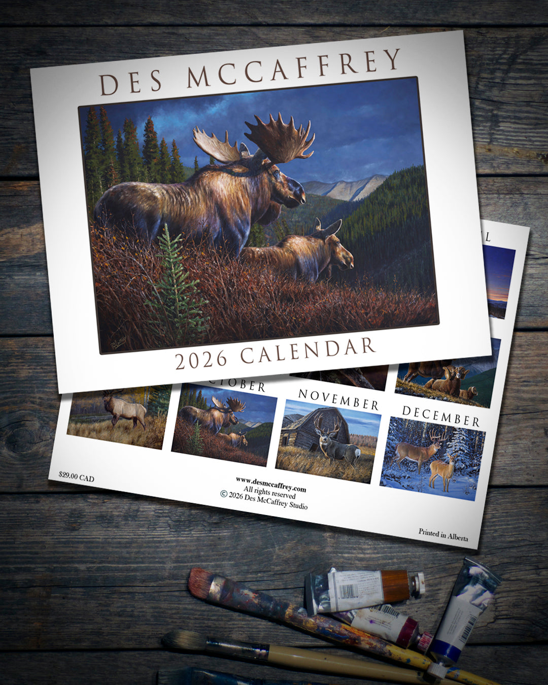 2026 wall calendar by wildlife artist Des McCaffrey featuring a cover illustration of two moose in an autumn mountain landscape, shown on a rustic wooden surface with paintbrushes and tubes of oil paint.
