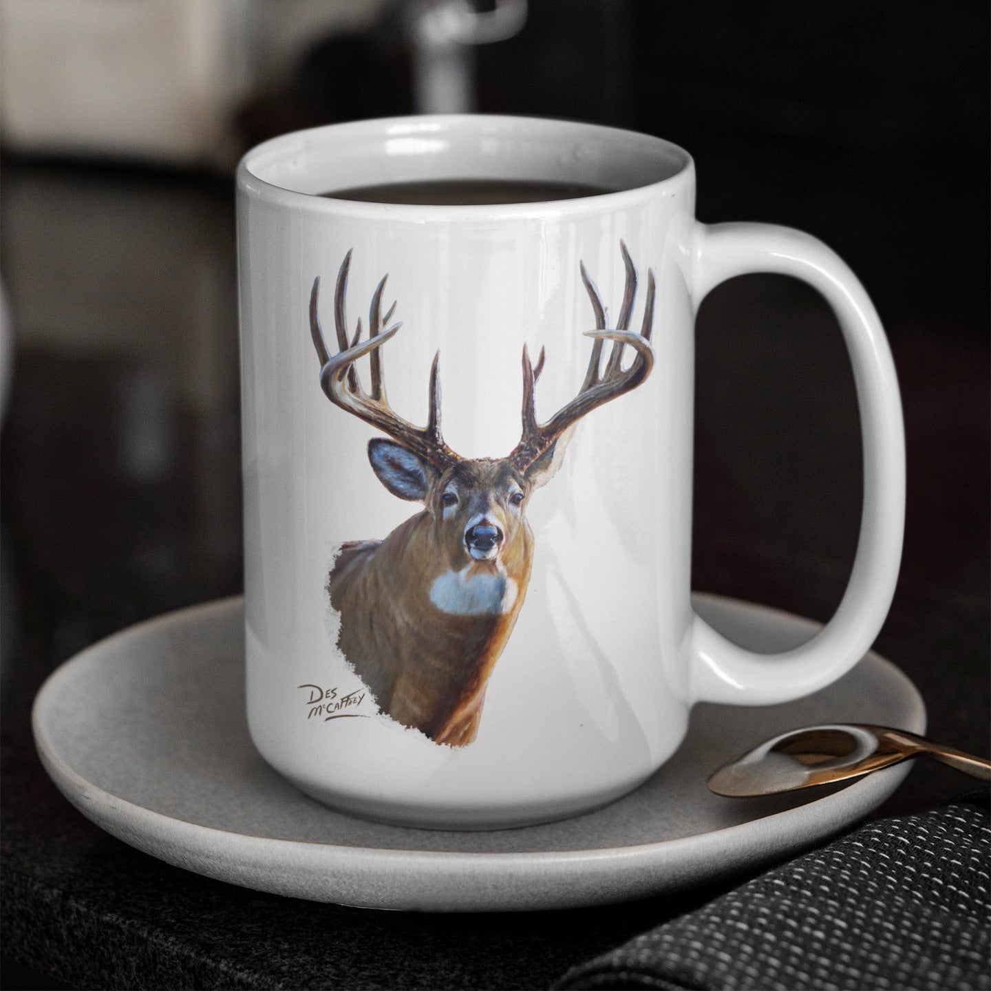 White ceramic coffee mug featuring a painting of a whitetail buck with large antlers, artwork by Canadian wildlife artist Des McCaffrey, resting on a saucer with spoon.
